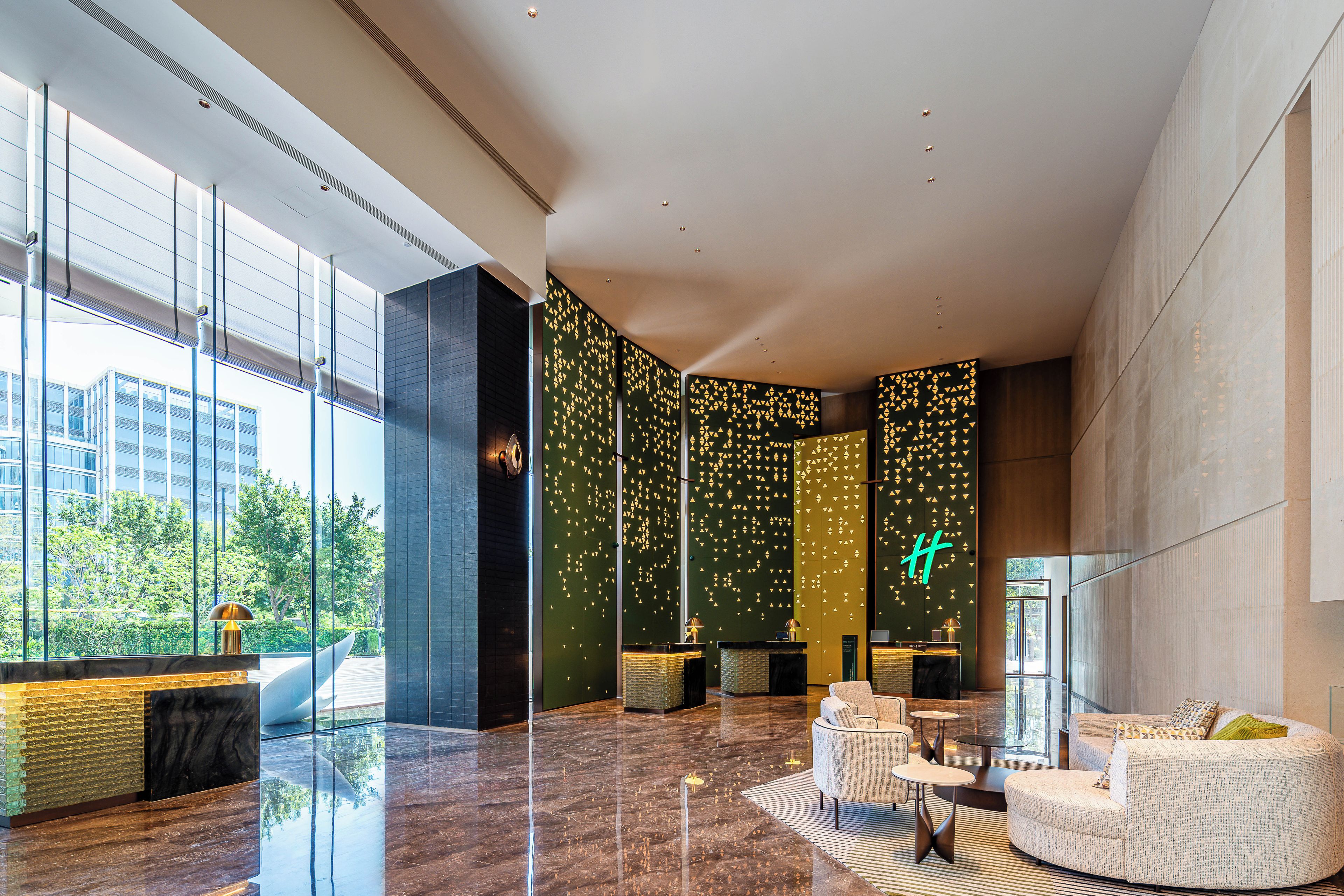 A modern lobby with a reception desk, seating area, and large windows offering views of greenery.