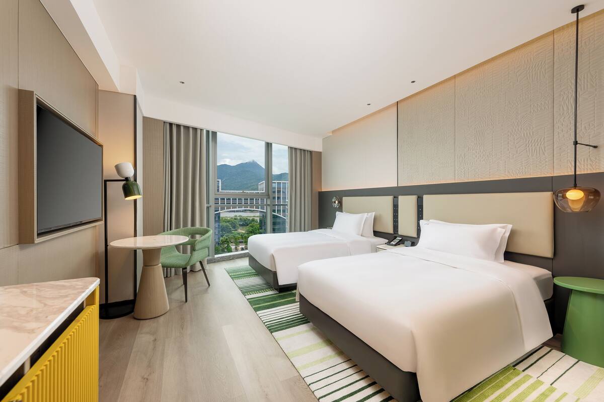 A hotel room with two beds, a TV, a small table, and a view of the cityscape.