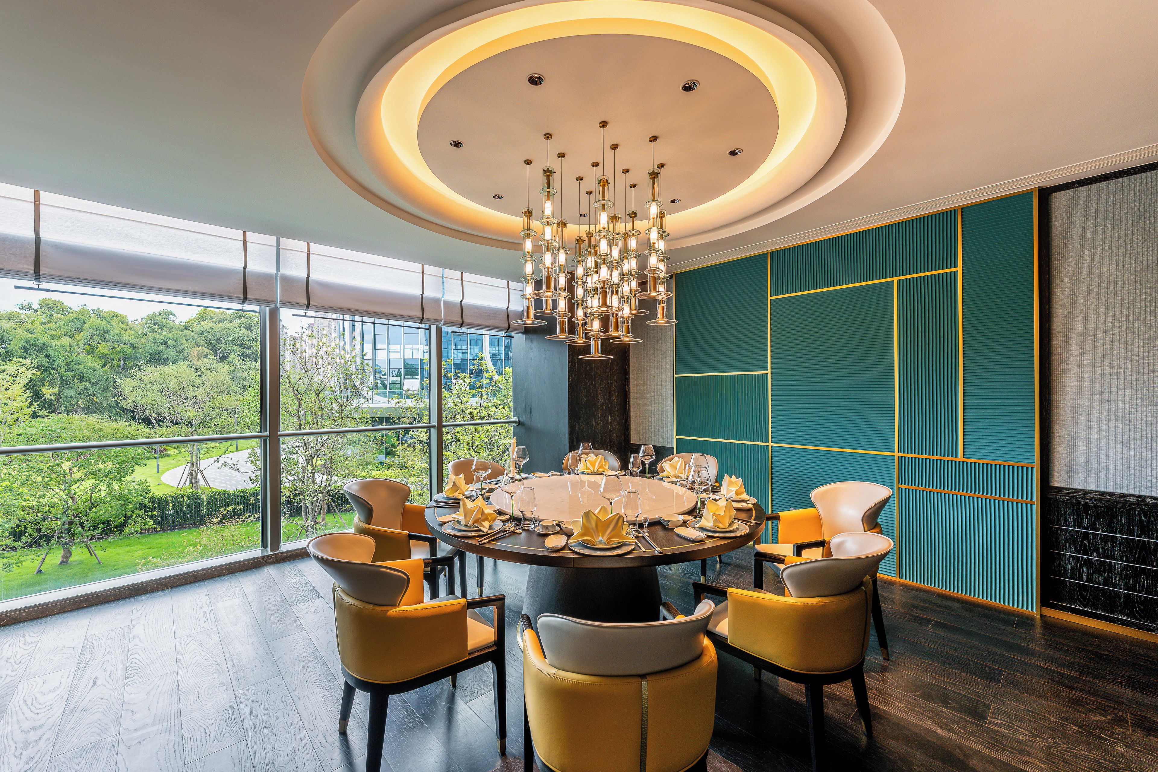 A modern dining area with a round table set for six, surrounded by yellow chairs, large windows offering a view of greenery, and a stylish ceiling light fixture.