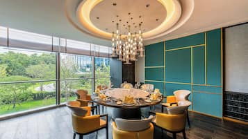 A modern dining area with a round table set for six, surrounded by yellow chairs, large windows offering a view of greenery, and a stylish ceiling light fixture.