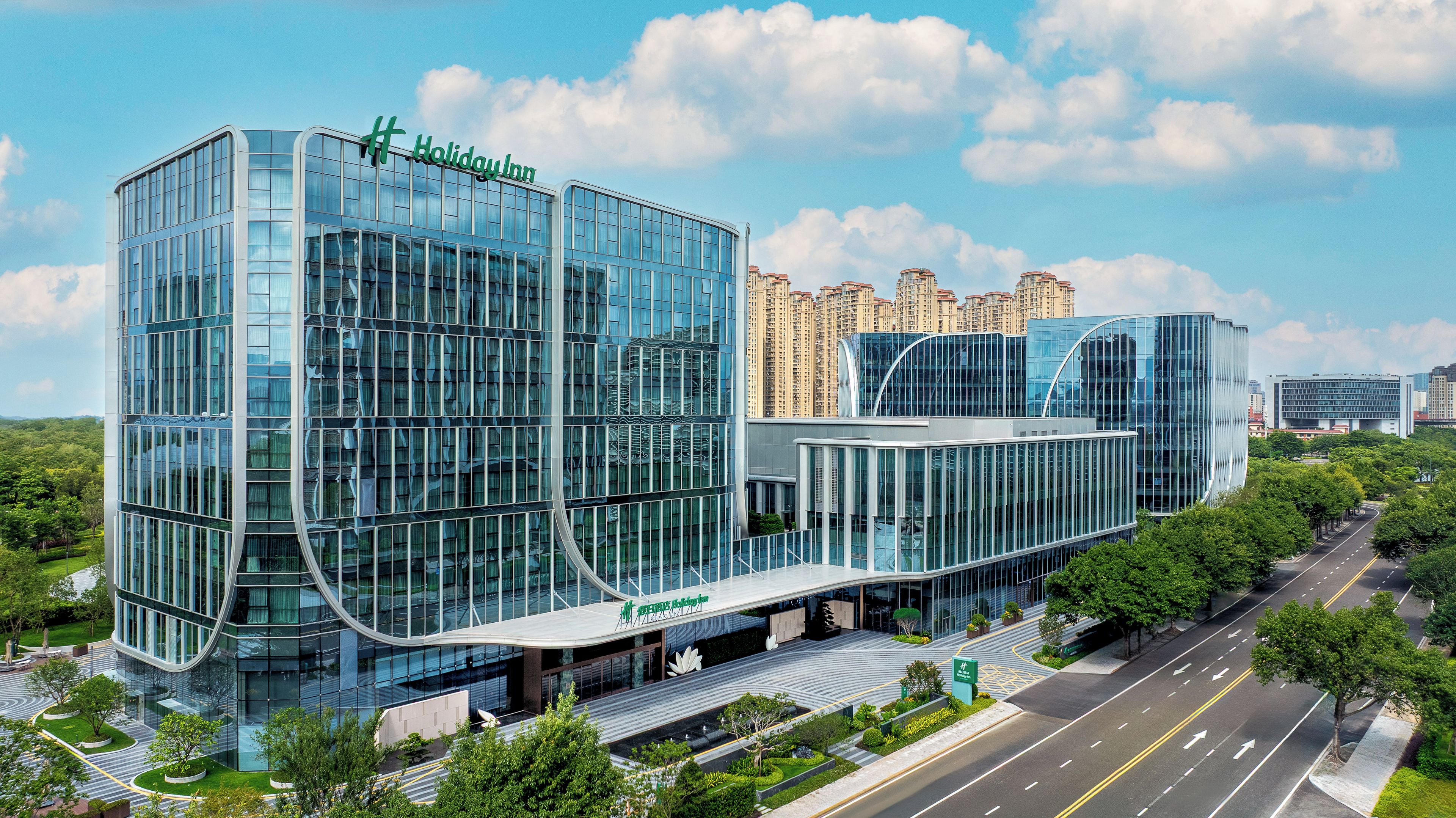 A modern Holiday Inn hotel with a glass facade, located in an urban area with surrounding high-rise buildings.