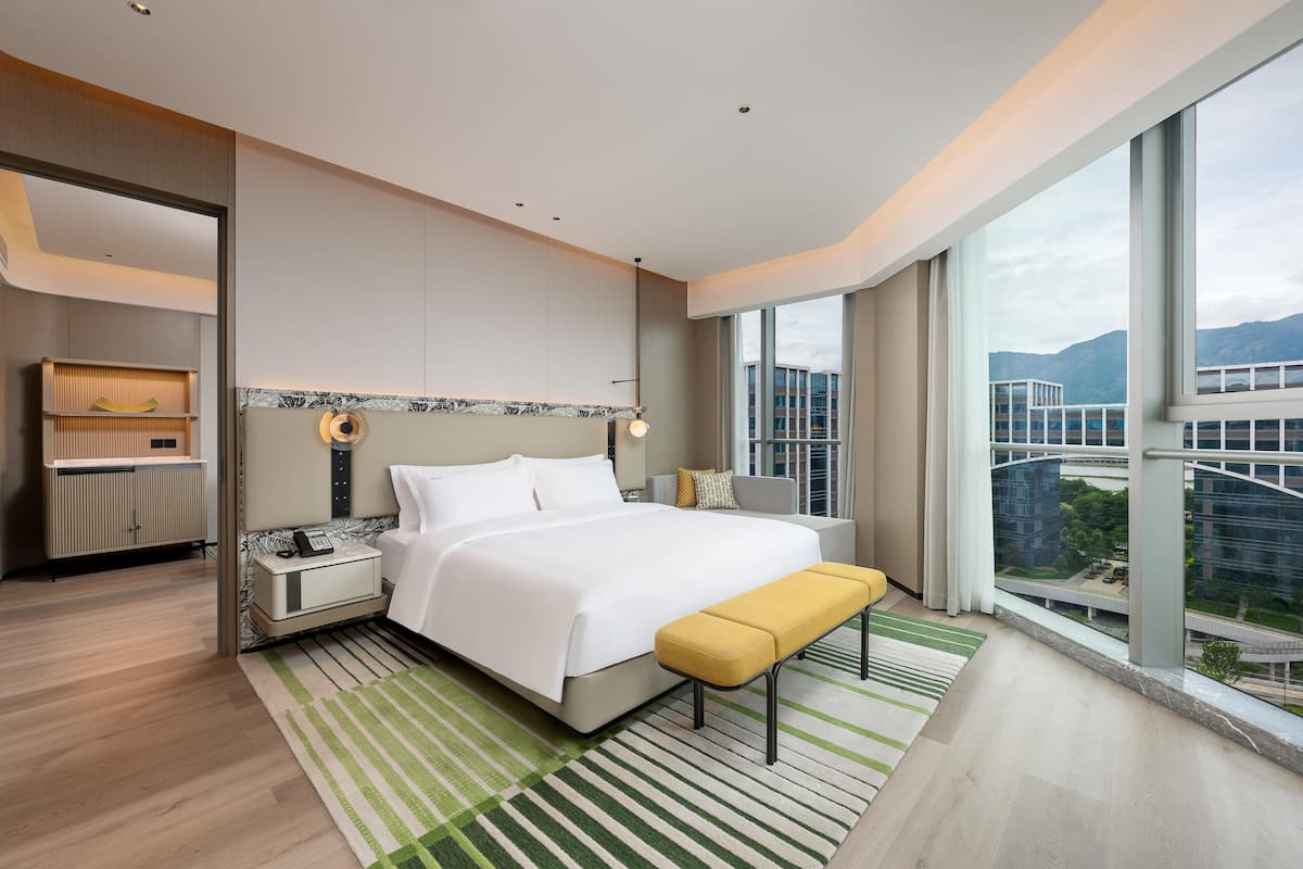 A modern hotel room with a large bed, a bedside table, a bench, a view of the city, and a large window.