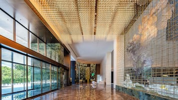 Um hall espaçoso com piso de mármore refletivo, grandes janelas com vista para áreas verdes e teto decorado com luminárias pendentes.