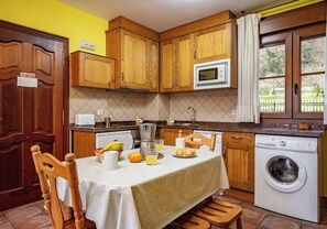 Een keuken met houten kastjes, een magnetron, een wasmachine en een eettafel gedekt met fruit en een koffiezetapparaat.