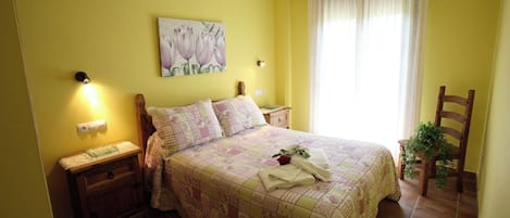 Un dormitorio con cama, mesitas de noche, lámpara de pared, un cuadro, ventana con cortinas y una silla.