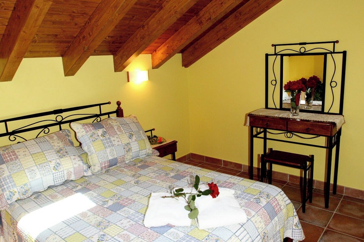 A bedroom with a wooden ceiling, a bed with a quilt, a bedside table with a lamp, and a mirror on the wall.