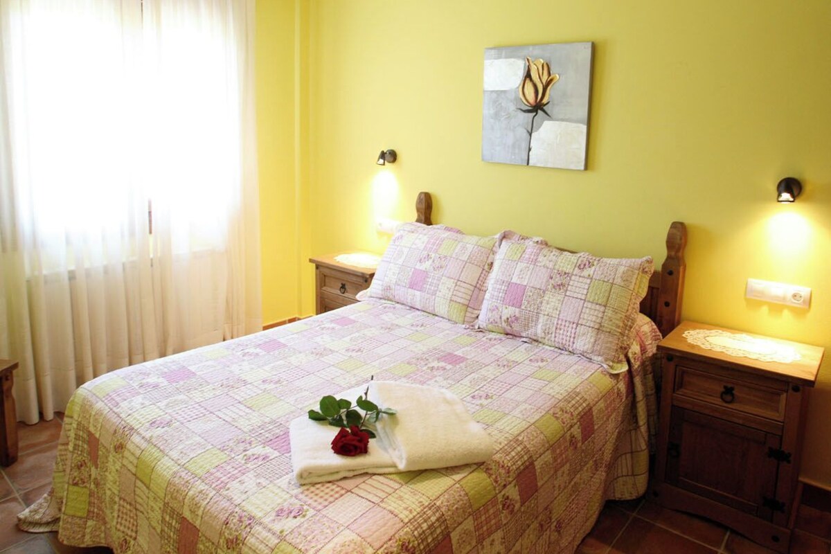 A bedroom with a large bed, two bedside tables, a nightstand, a ceiling light fixture, a wall-mounted artwork, and a window with curtains.