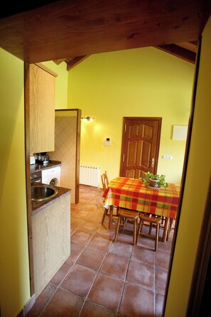 A kitchen with a built-in sink, a coffee machine, and a washing machine. There is a dining area with a table covered by a plaid tablecloth and wooden chairs. A wooden door and a light switch are visible on the wall. - Apartamentos Rurales La Viña (Cangas de Onis)