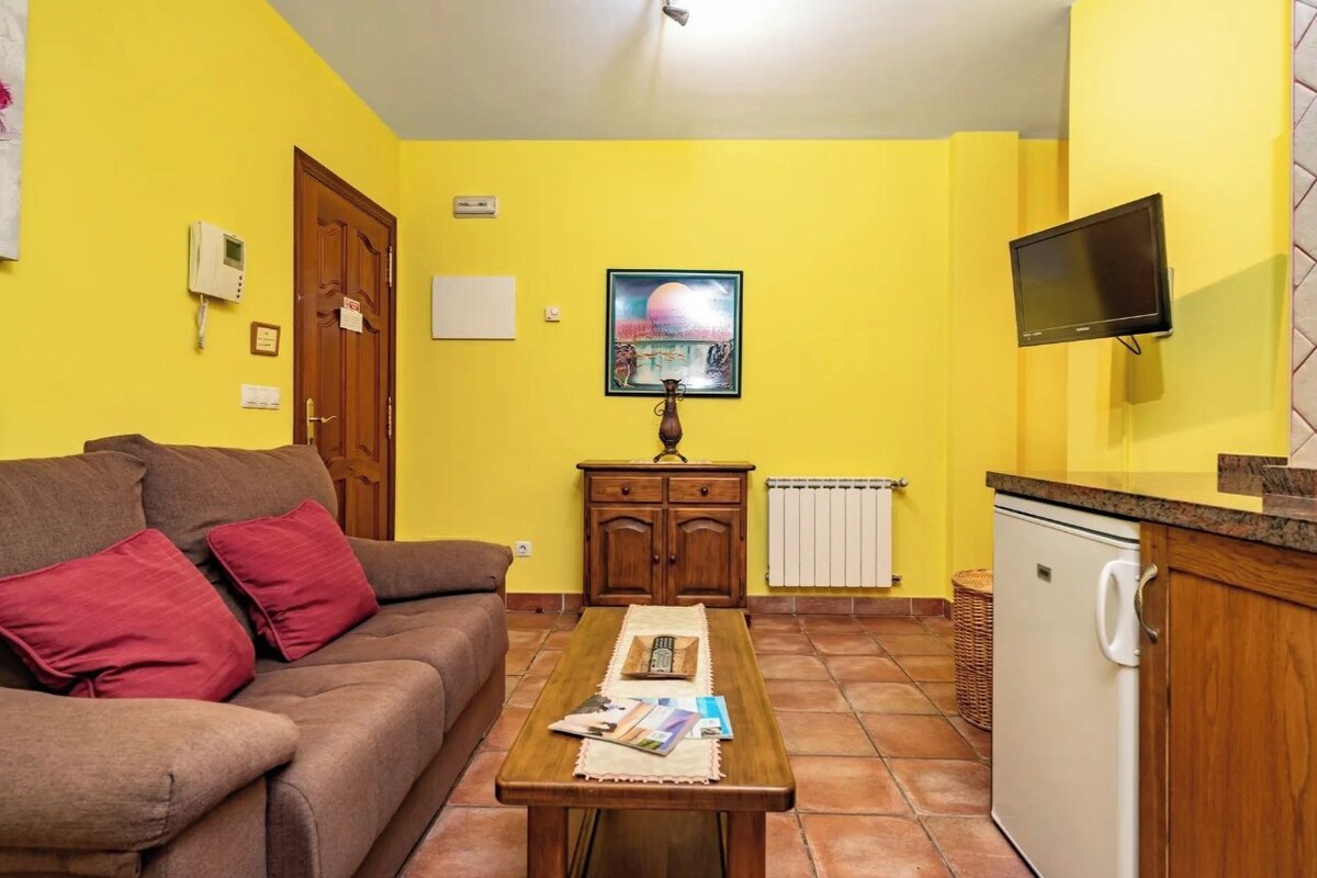 A living room with a sofa, a wooden table with magazines, a painting on the wall, a television, and a kitchen area with a refrigerator.