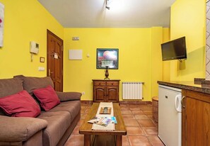 A living room with a sofa, a wooden table with magazines, a painting on the wall, a television, and a kitchen area with a refrigerator. - Apartamentos Rurales La Viña (Cangas de Onis)
