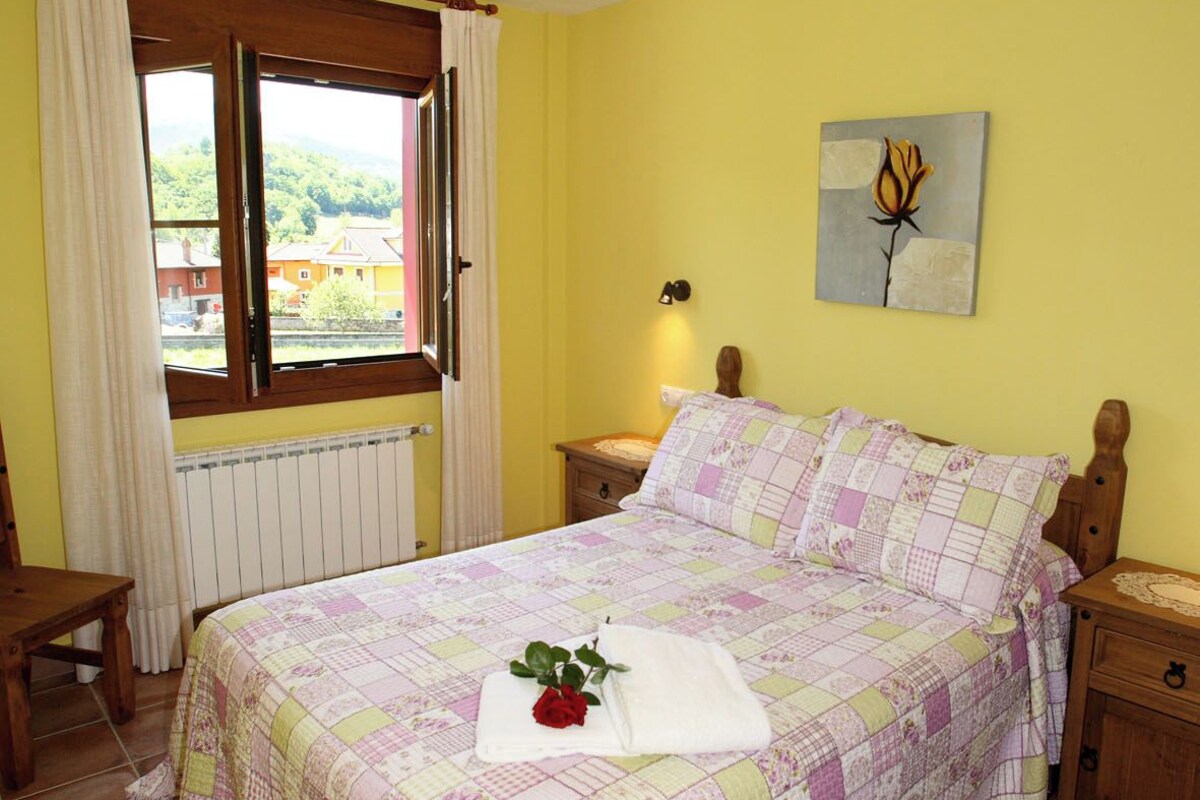 A bedroom with a bed, a nightstand, a window with curtains, a radiator, and a wall-mounted light fixture.