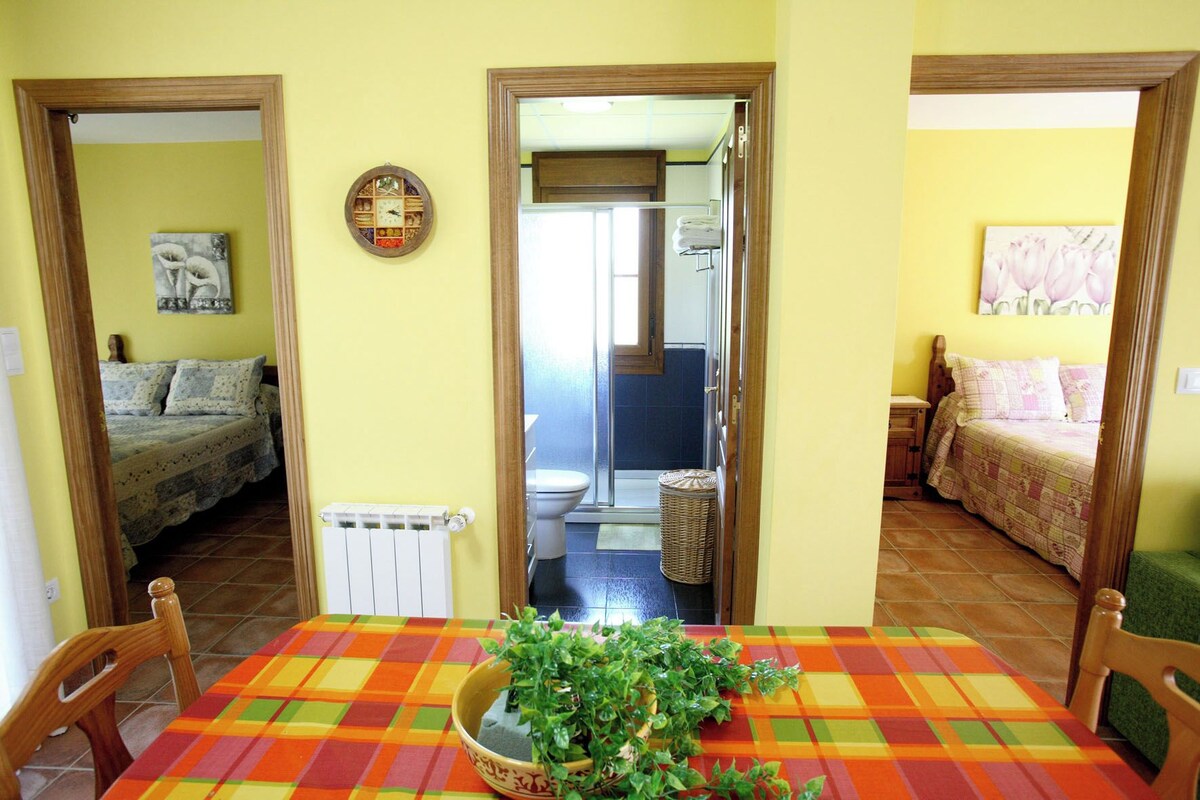 A room with a bed, a table with a plant, and a bathroom visible through an open door.