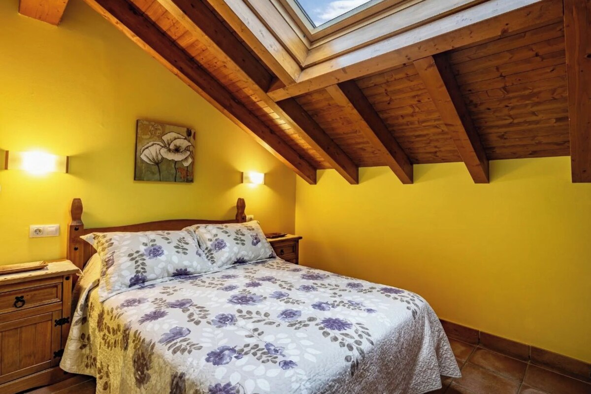 A bedroom with a skylight, wooden beams, a floral bedspread, and a painting on the wall.