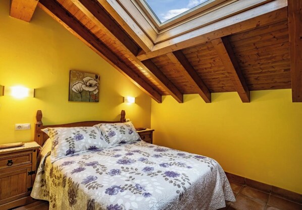 A bedroom with a skylight, wooden beams, a floral bedspread, and a painting on the wall.