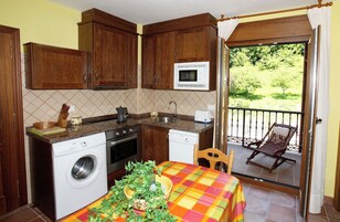 Cocina compacta con lavadora, microondas y lavaplatos. Hay un comedor con mesa y sillas, y un balcón con una silla.