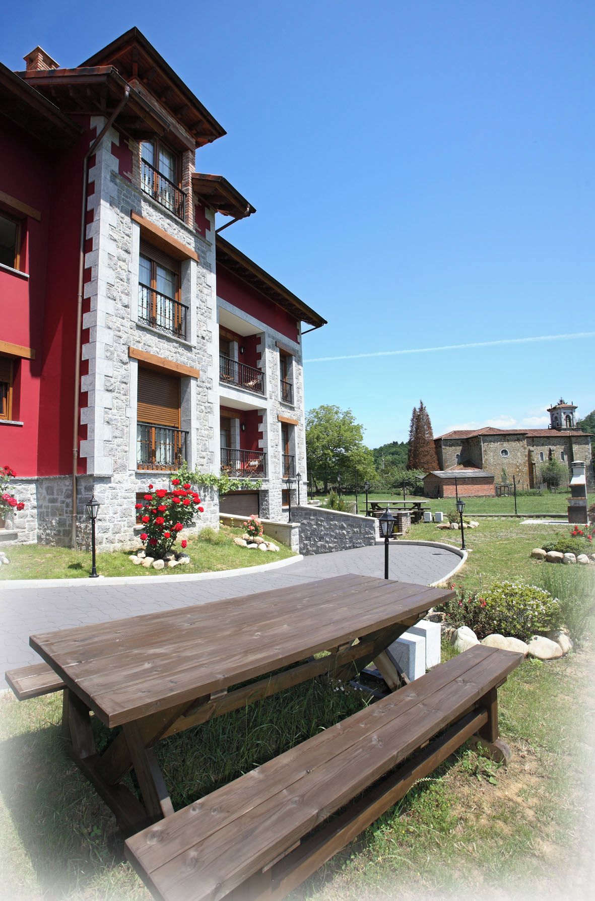A multi-story building with stone and red exterior walls, a balcony, and a paved area with picnic tables.