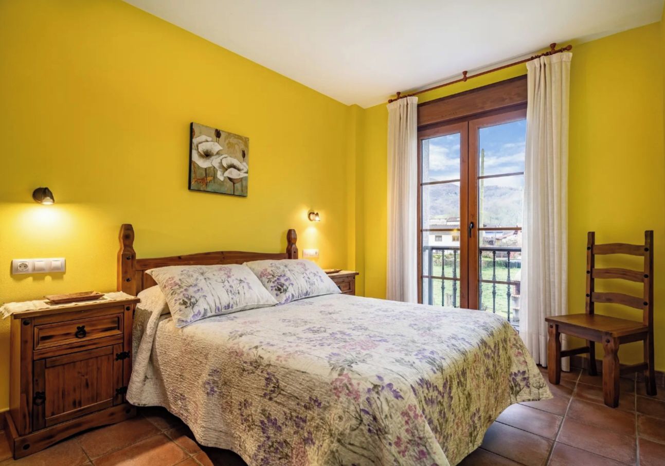A bedroom with a bed, bedside tables, a chair, a window with curtains, and a wall-mounted light fixture.