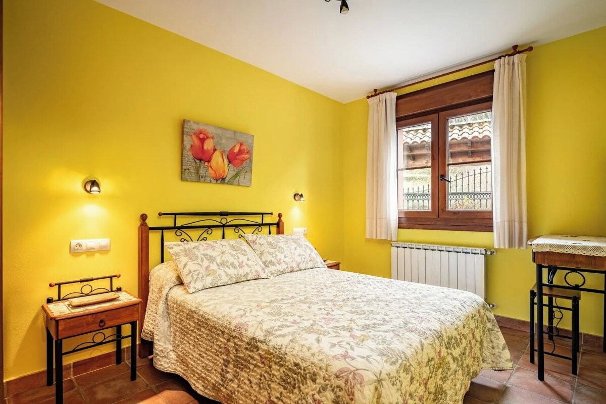 A bedroom with a bed, a nightstand, a table, a window with curtains, and a wall painting.