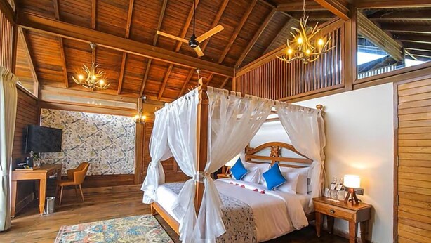 A four-poster canopy bed with a wooden headboard, a bedside table with a lamp, and a patterned rug on the floor. - Toyam By Orchid Hotels (Bhor)