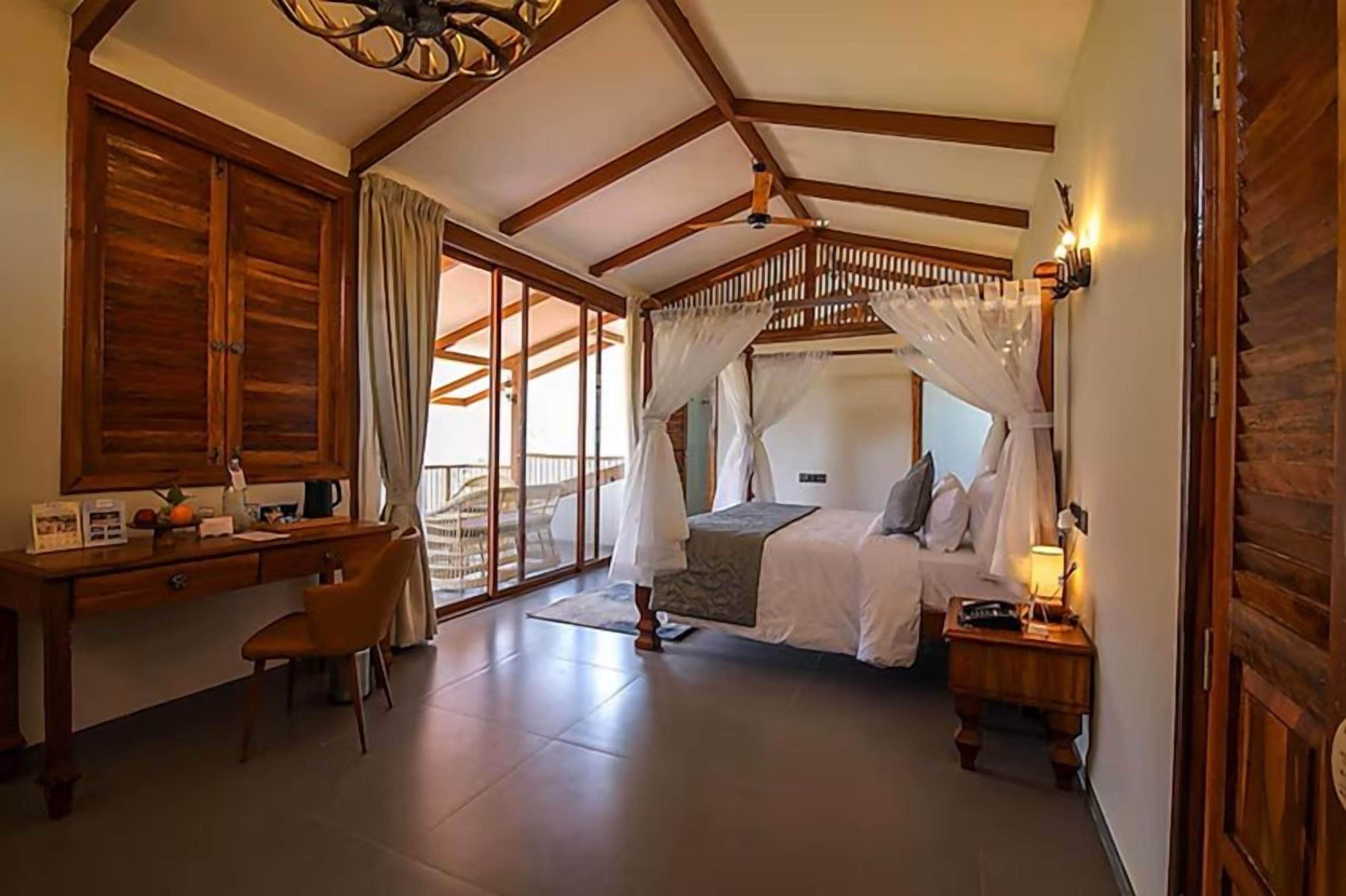A bedroom with a bed, a desk, and a chair. There is a window with curtains and a view of the outside.