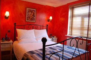 Une chambre avec un lit, une table de chevet, une lampe fixée au mur et une fenêtre avec des stores.