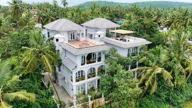 A multi-story white building with a flat roof, surrounded by dense greenery and palm trees. - Elivaas Vacasa - Goa (Candolim)