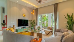 A modern living room with a flat-screen TV, comfortable seating, a coffee table, and a large window with a view of greenery. - Elivaas Vacasa - Goa (Candolim)