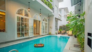 A swimming pool with a floating fruit basket, a covered patio with chairs, and a building with arched windows. - Elivaas Vacasa - Goa (Candolim)