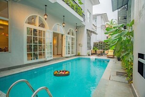 A swimming pool with a floating fruit basket, a covered patio with chairs, and a building with arched windows. - Elivaas Vacasa - Goa (Candolim)