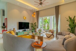 A modern living room with a flat-screen TV, comfortable seating, a coffee table, and a large window with a view of greenery. - Elivaas Vacasa - Goa (Candolim)
