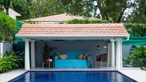 Una piscina con zona de estar cubierta, techo con baldosas y follaje circundante.