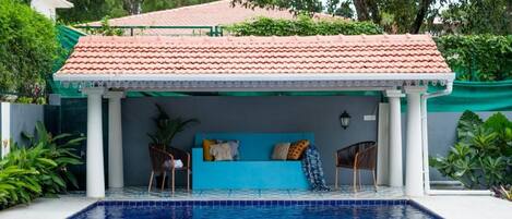 A pool with a covered seating area, a tiled roof, and surrounding greenery.
