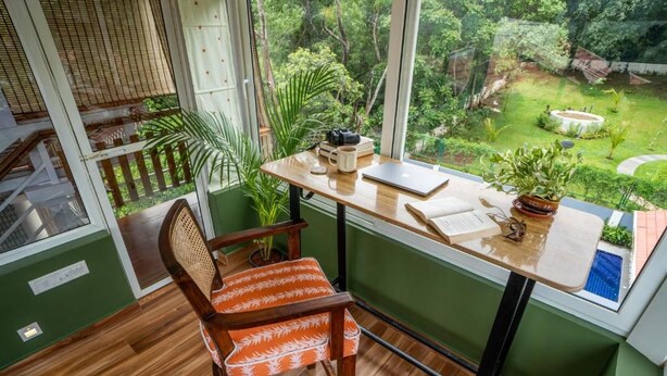 A room with a wooden desk, a chair, a window with a view of a garden, and a small table with a plant. - Elivaas Bludoor - Goa (Assagao)