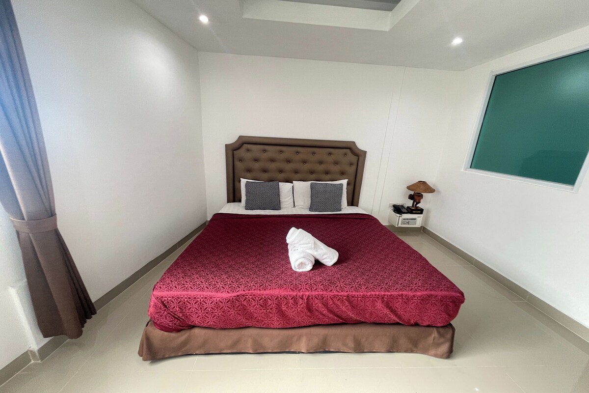 A neatly made bed with a red patterned bedspread and a folded towel, a bedside lamp, and a window with a green view.