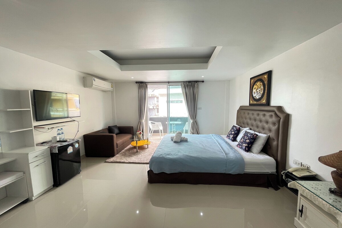 A modern hotel room with a bed, a TV, a desk, and a small kitchenette.