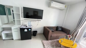 A modern living room with a flat-screen TV, a white desk with drawers, a black mini-fridge, a brown sofa, and a glass coffee table. - W-9 Hotel By Zuzu (Pattaya)