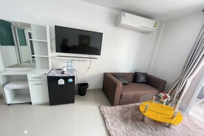 A modern living room with a flat-screen TV, a white desk with drawers, a black mini-fridge, a brown sofa, and a glass coffee table. - W-9 Hotel By Zuzu (Pattaya)