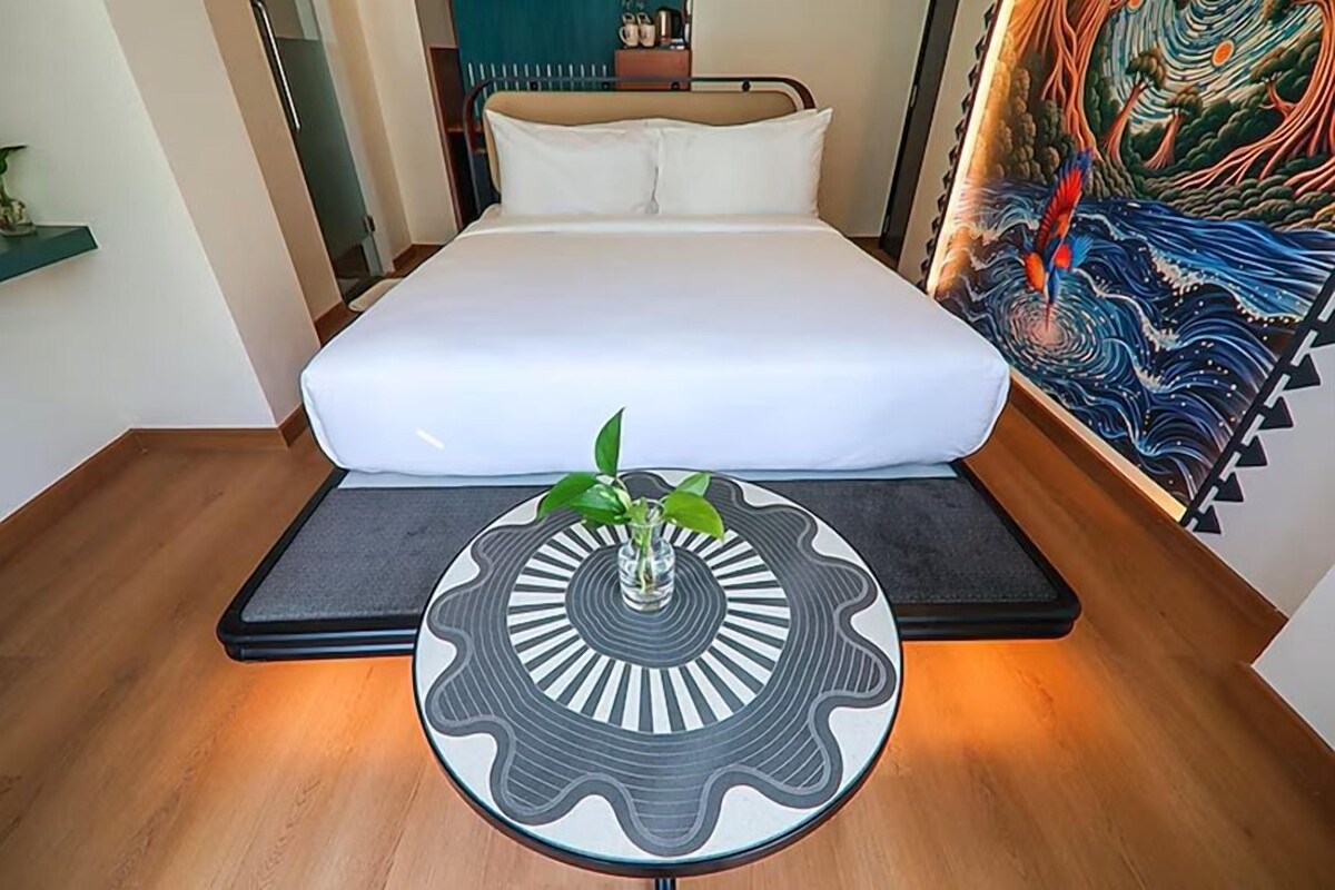 A neatly made bed with a white mattress and pillows, a small table with a decorative vase, and a large painting on the wall.