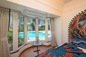 A room with a large window, a bed, a small table, and a colorful tapestry.