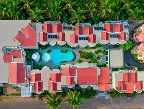Una vista aerea di un resort con diversi edifici a tetti rossi, una piscina centrale e vegetazione circostante.