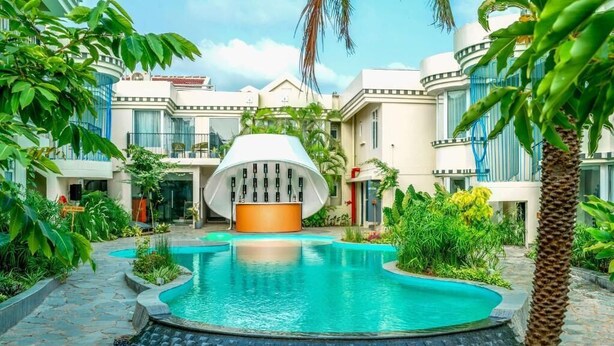 A pool area with a modern building in the background, surrounded by lush greenery. - The Orchid Passaros (Benaulim)