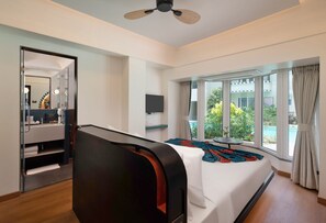 A hotel room with a bed, a ceiling fan, a TV, a window with a view of greenery and a building, and a bathroom with a mirror and towel rack. - The Orchid Passaros (Benaulim)