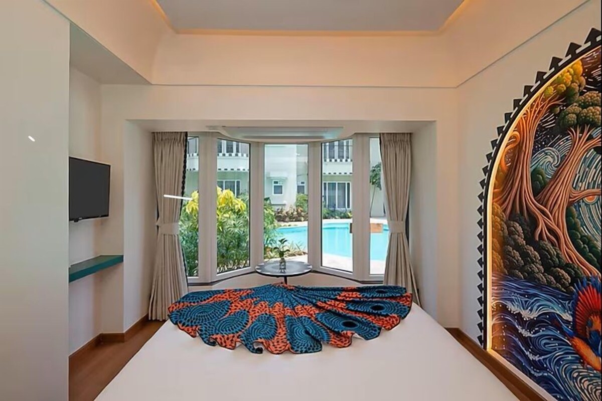 A hotel room with a large bed, a TV, a view of a swimming pool, and a large wall painting.