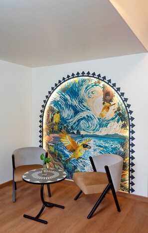 A room with a mural of a bird and waves, two chairs, a small table with a vase, and a wooden floor. - The Orchid Passaros (Benaulim)