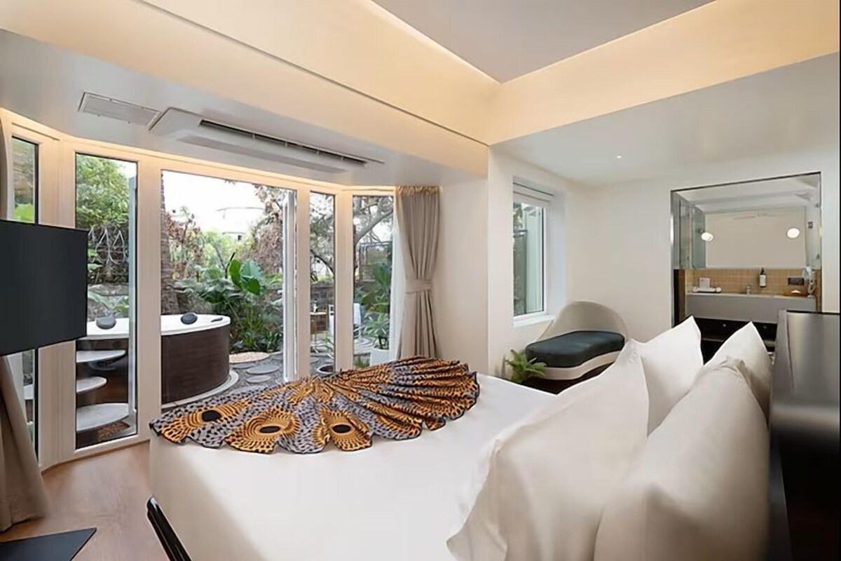 A modern hotel room with a large bed, a sitting area with a view of the garden, and a separate bathroom with a mirror.