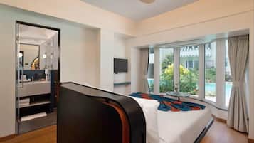 A hotel room with a bed, a TV, a ceiling fan, a window with a view of greenery and a building, and a small table with a lamp.