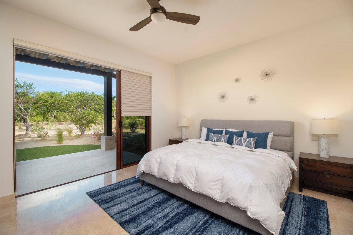 A bedroom with a bed, a ceiling fan, a nightstand, and a view of a garden through a sliding door.