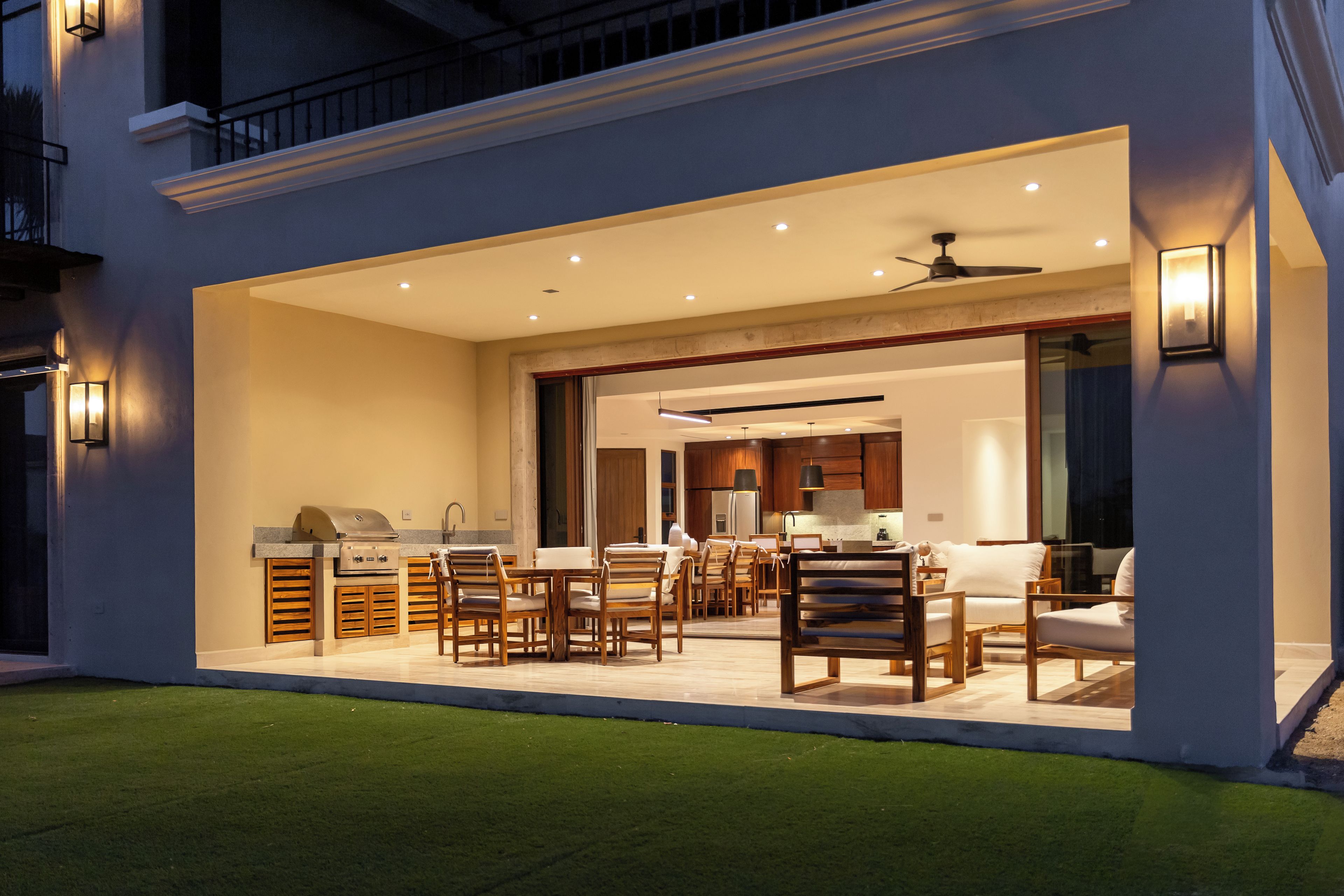 An outdoor dining area with wooden furniture and a grill, connected to a kitchen with modern appliances.