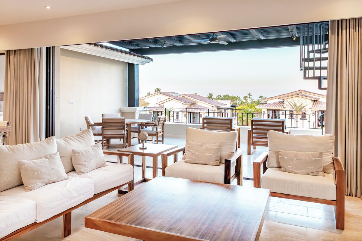 A modern lounge area with a sofa, chairs, and a coffee table. There is a view of a building with a tiled roof and palm trees in the background.