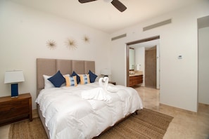 Une chambre d’hôtel avec un grand lit, une table de chevet en bois, une lampe fixée au mur, des appliques décoratives et une salle de bain visible à travers une porte ouverte.